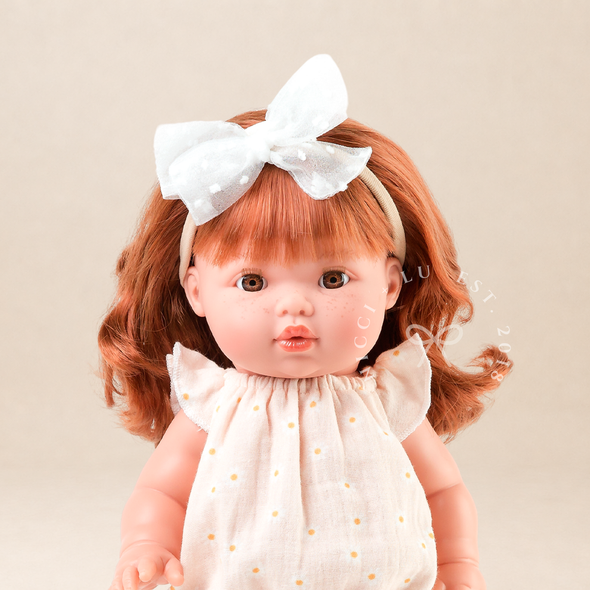 Portrait of 34cm Mini Colettos Sophia wearing the Willow Muslin Romper in Daisy Print with a white bow headband.