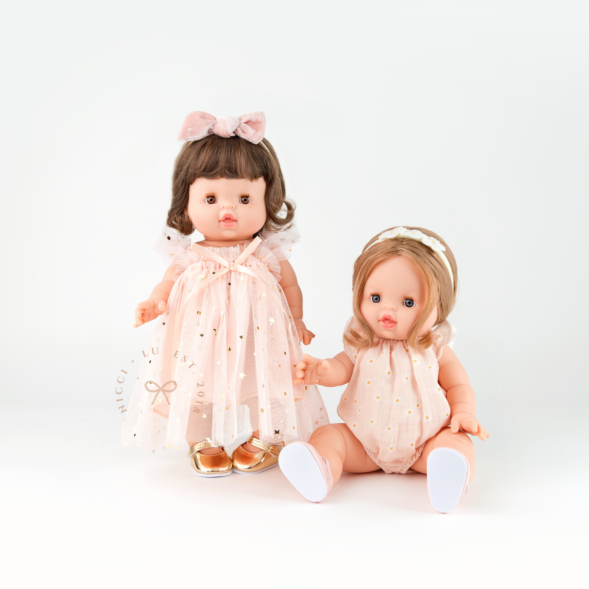 Two Minikane dolls wearing the Rose Blush Tulle Ballet Dress and the Willow Muslin Romper in Daisy Print.