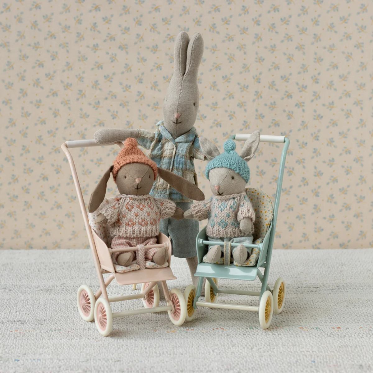 Maileg winter Rabbit Micro Sand and Mint sitting in a strollers with a larger plush rabbit pushing them, against a floral wallpaper background.