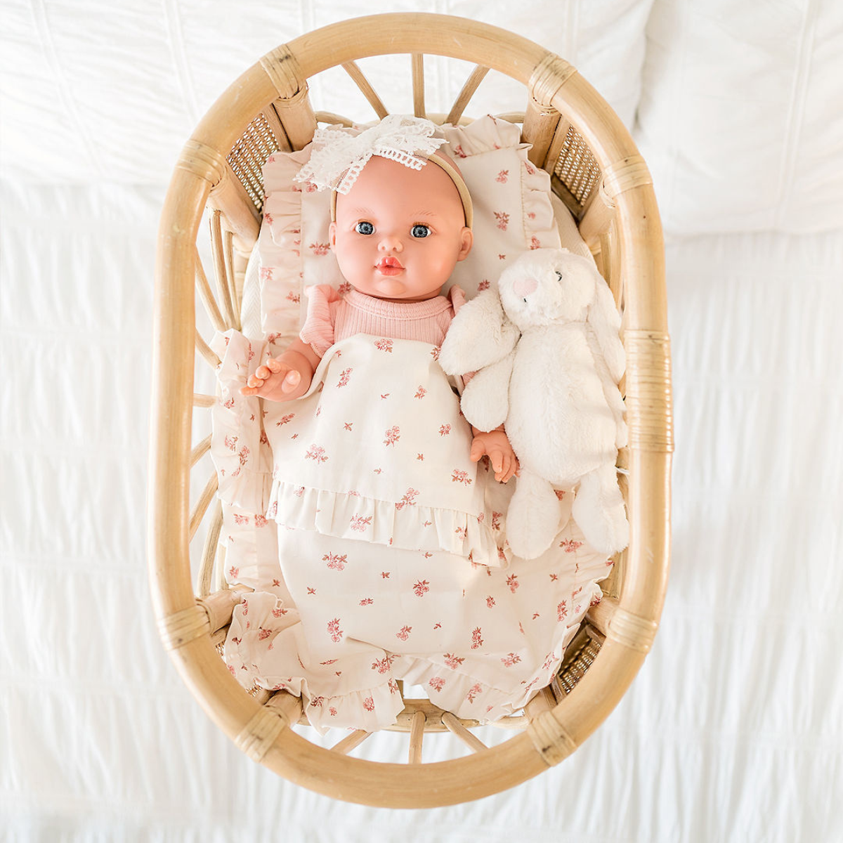 Mini Coletto Mila 34cm doll in a Doll Clothes lying on a Nicci + Lu comforter with a rattan bassinet.