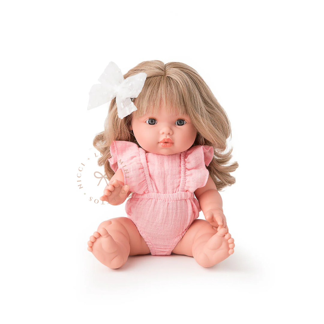 Kate looks adorable wearing a Muslin Flutter Romper in  pink with delicate ruffle detailing and a white hair bow. Her beautiful blonde curls complete the look, showcasing the 34cm Mini Colettos doll dressed in a sweet pastel outfit.