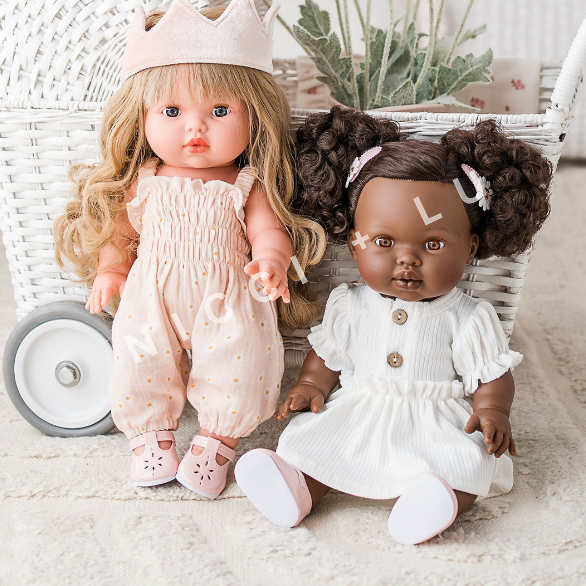 Lyla and Jedda are Mini Coletto dolls and they are posing next to a stroller in their Little Mary Jane shoes, Smocked Daisy Jumpsuit outfit , Ivory Puff sleeve dress and velvet crown doll accessory.