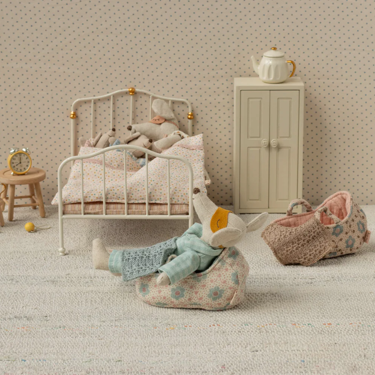 The Maileg Baby Mouse Carry Cot in rose, perfect for transporting tiny mouse dolls during play. Miniature bedroom scene with a bed, cabinet, and toys on a textured floor.
