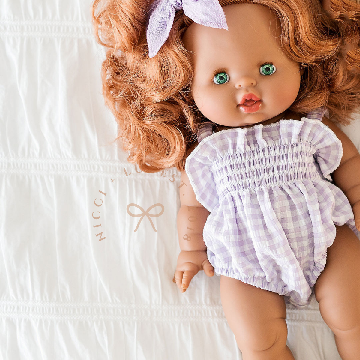 Minikane Juliette doll wearing the Olivia Smocked Bubble Romper in Lilac Gingham while lying down.