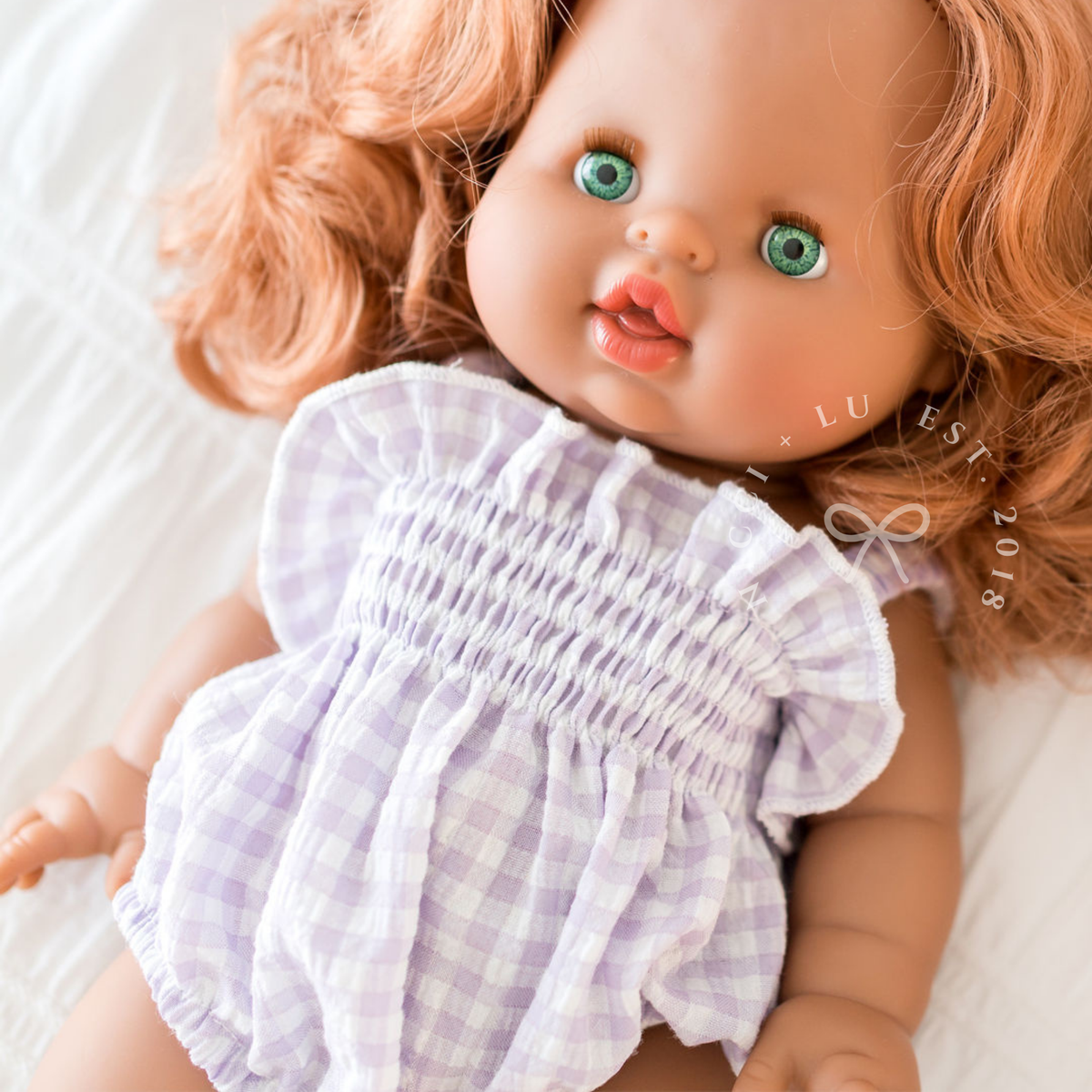 Mini Colettos Isabel doll lying down wearing the Olivia Smocked Bubble Romper in Lilac Gingham.