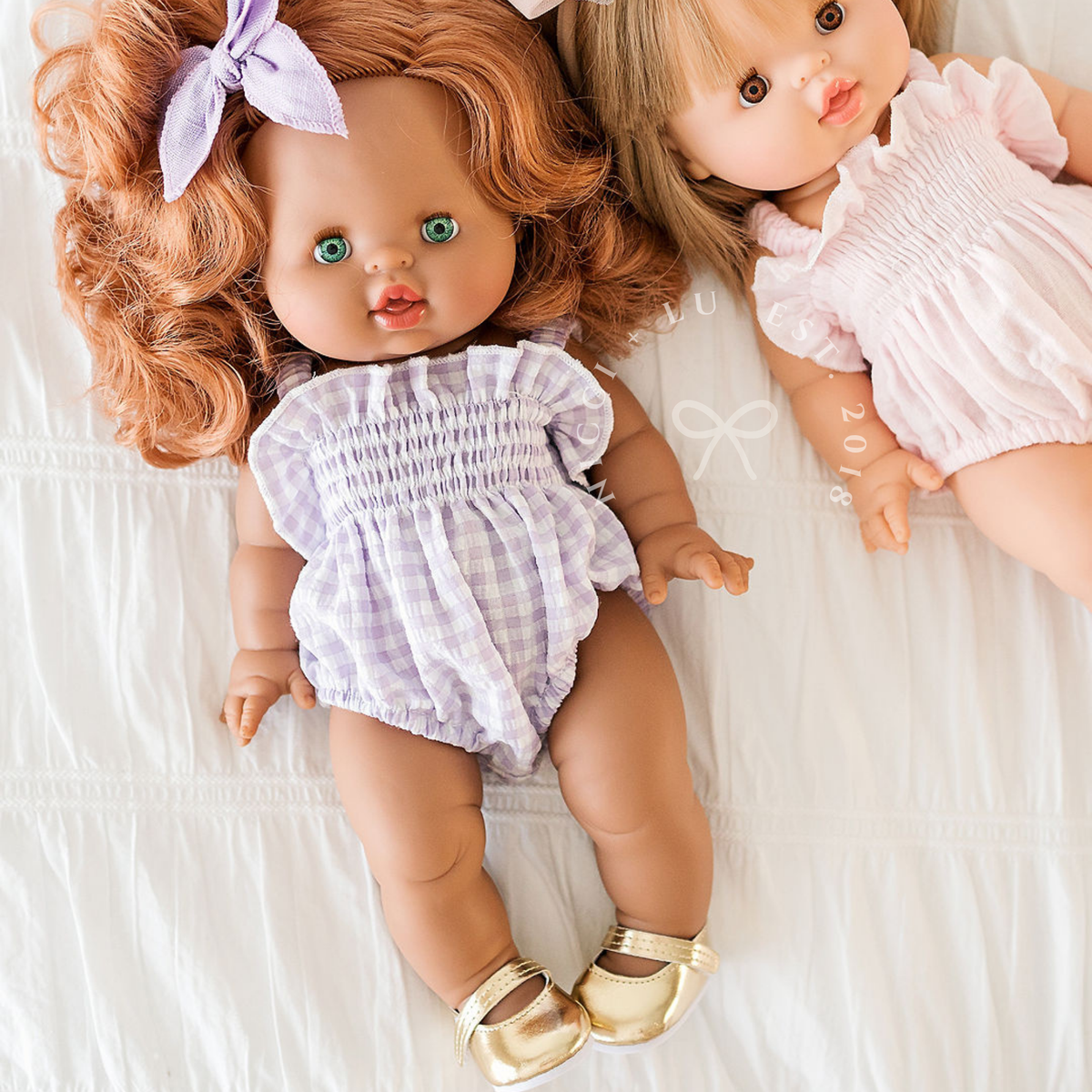 Minikane Juliette doll wearing the Olivia Smocked Bubble Romper in Lilac Gingham lying beside another doll.